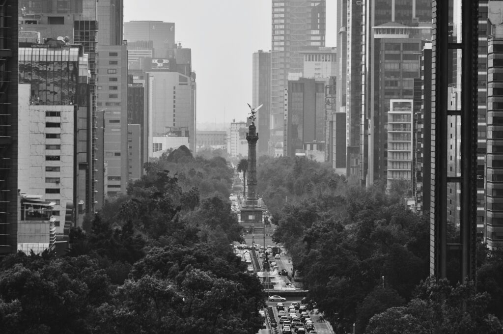 Fotografía en blanco y negro del icónico Ángel de la Independencia en Ciudad de México, ubicado en Paseo de la Reforma, rodeado por rascacielos y edificios modernos, con tráfico y árboles alineados en la avenida.